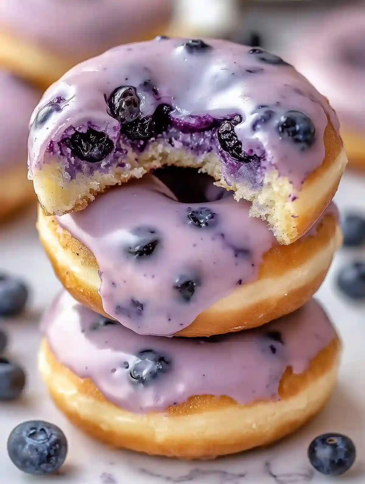 Stacked blueberry-glazed donuts with a bite, blueberries, and marble background.