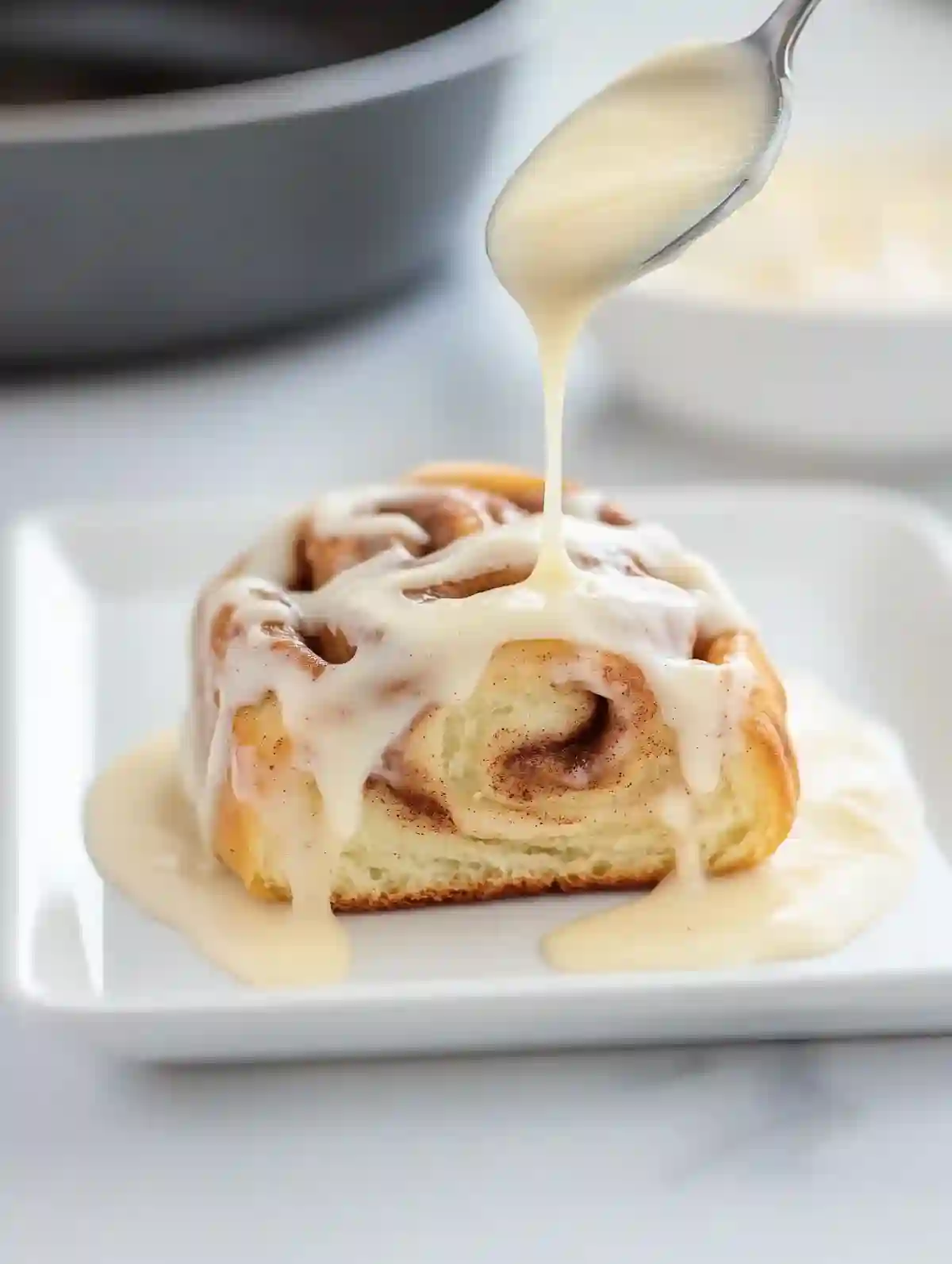Cinnamon roll on white plate being drizzled with icing, soft lighting.