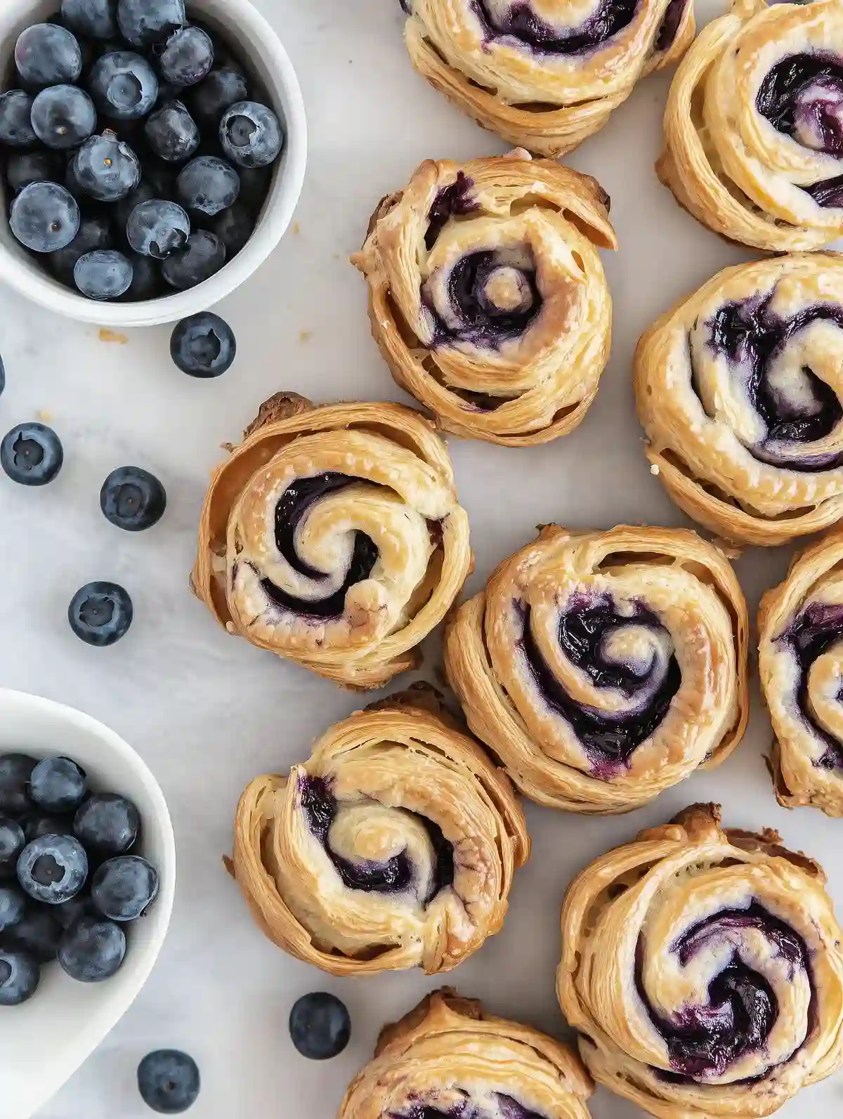Golden blueberry cruffins arranged in a circle on marble, with blueberries nearby.
