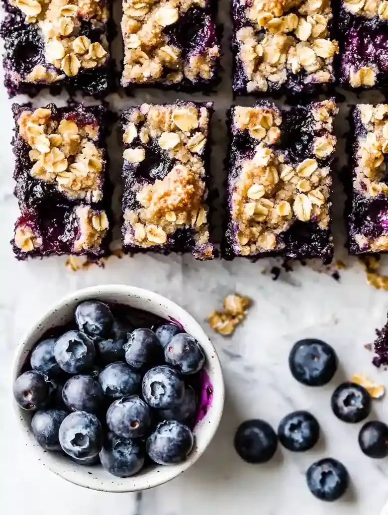 Blueberry oat bars with golden topping and fresh blueberries on marble.