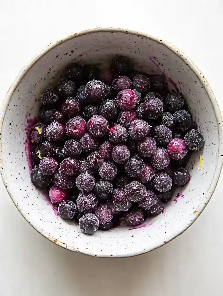 Bowl of frozen blueberries dusted with sugar and lemon zest on marble.