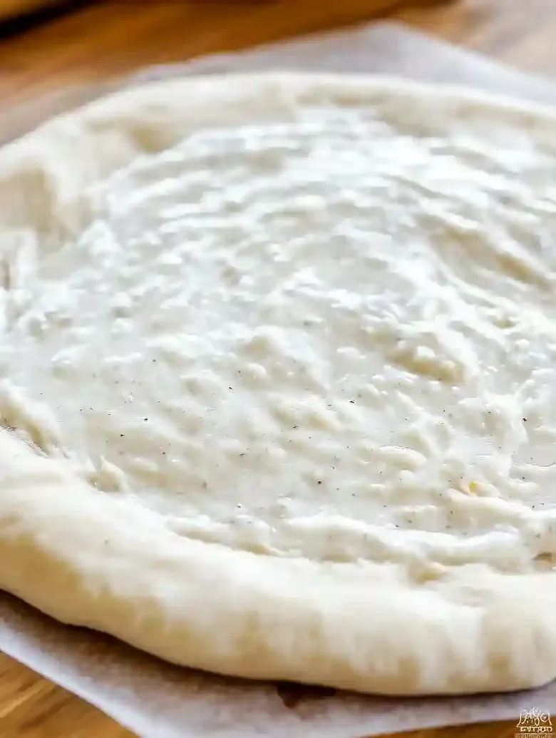 Creamy garlic pizza sauce spread on rolled-out dough on marble surface.
