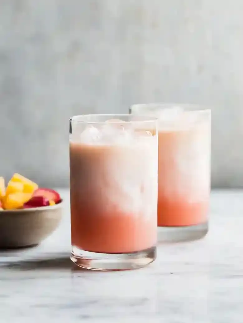 Tall glasses of gradient drink with ice cubes on marble surface, fruit bowl in background.