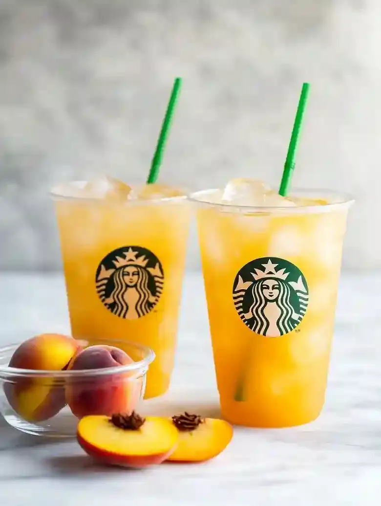 Two iced beverages with green straws beside fresh peach slices on marble.
