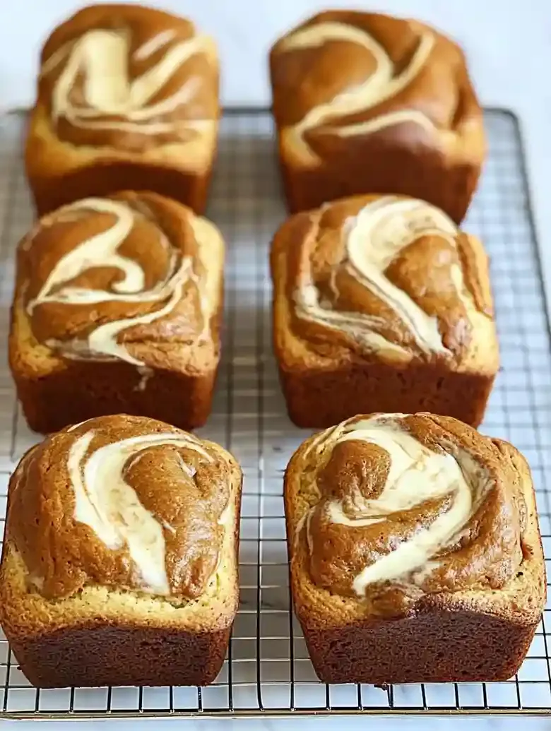 Ten mini pumpkin cream cheese loaves on a wire rack against marble.