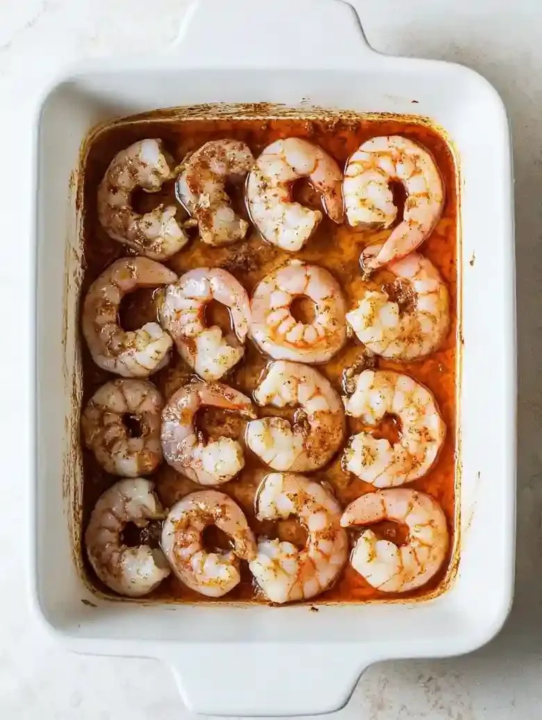 Raw shrimp in a baking dish, coated in marinade on a marble surface.