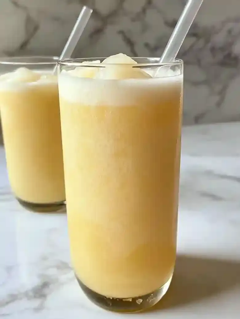 Tall glass of pineapple-coconut drink with frothy top on marble surface.