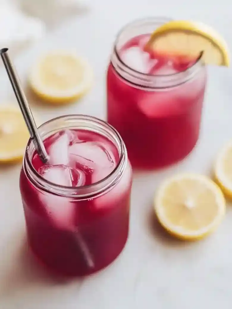 Two jars with vibrant drinks, ice, and lemon slices on marble surface.