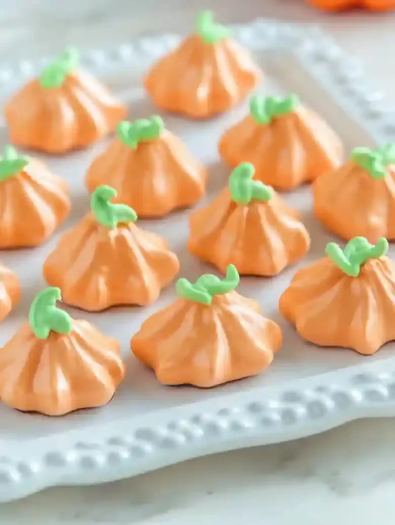 Pumpkin-shaped meringue cookies in orange with green stems on platter.