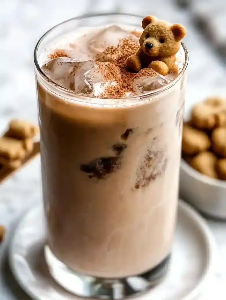 Iced latte with cinnamon and teddy bear-shaped cookie on marble surface.