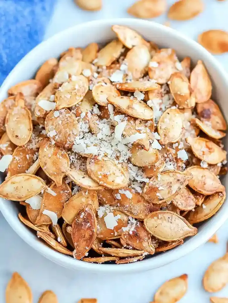 Bowl of garlic Parmesan pumpkin seeds on a marble surface, surrounded by seeds.