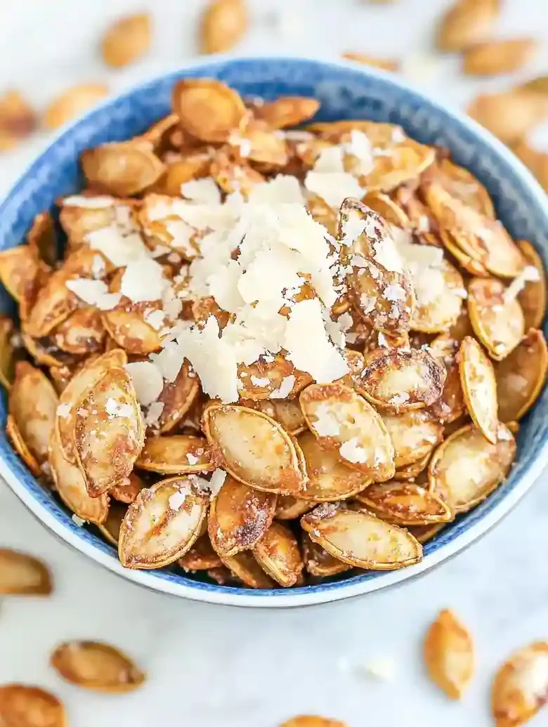 Bowl of garlic Parmesan pumpkin seeds on white marble surface.